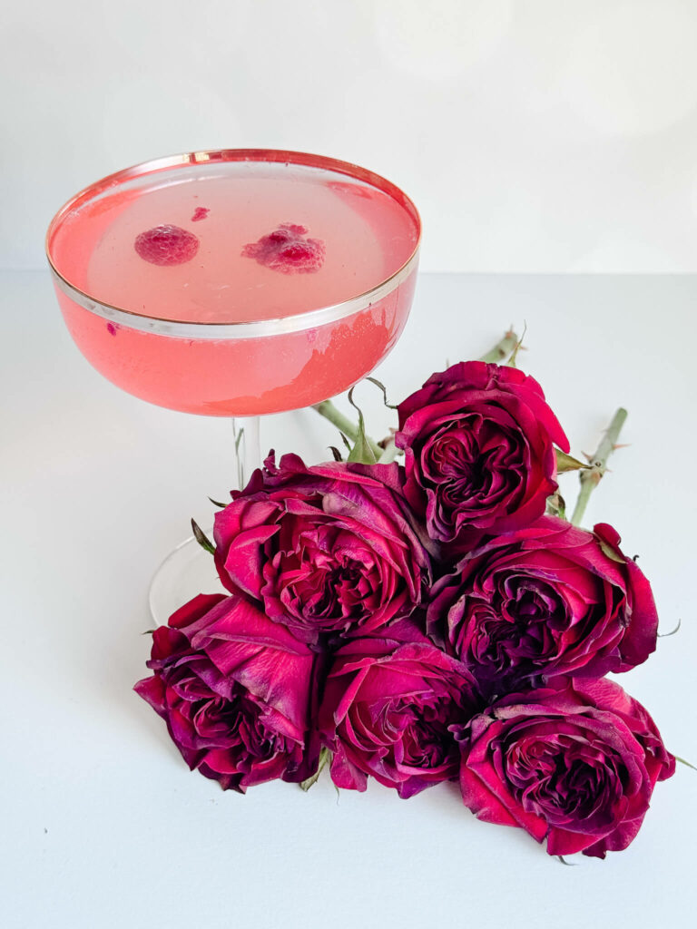 In a champagne coupe behind a bed of roses is the Kentucky Derby inspired cocktail Raspberry Lemonade Fizz garnished with 2 frozen raspberries.