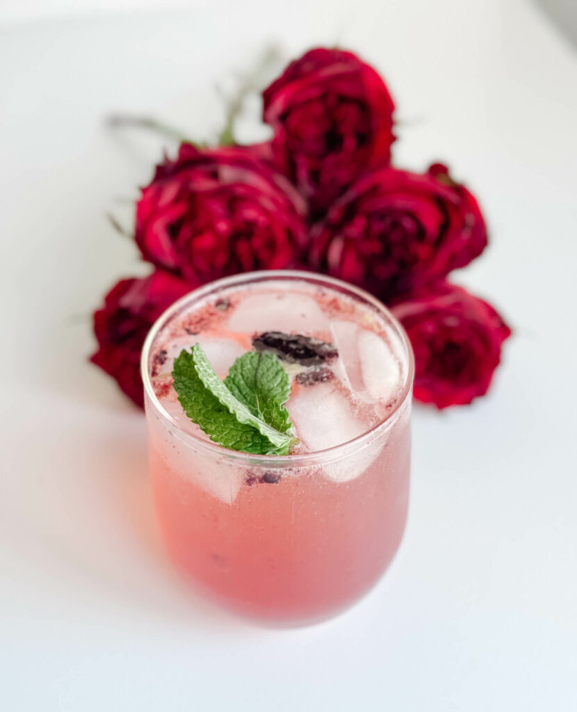 In a rocks glass full of ice in front of a bed of roses is a blackberry mint julep cocktail garnished with 2 sprigs of mint.