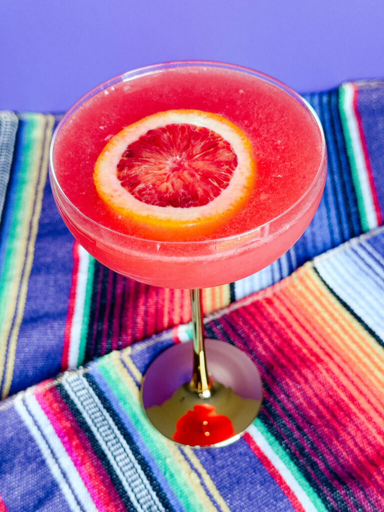 A bubbly amaretto blood orange margarita in a champagne coupe garnished with a blood orange wheel.