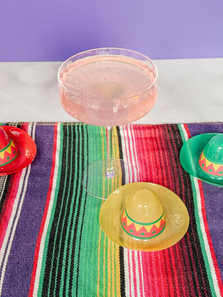 A sparkling pink lemonade margarita in a champagne coupe surrounded by mini sombreros.