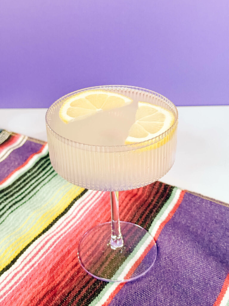 A sparkling sicilian lemonade margarita in a purple champagne coupe garnished with two lemon halves.